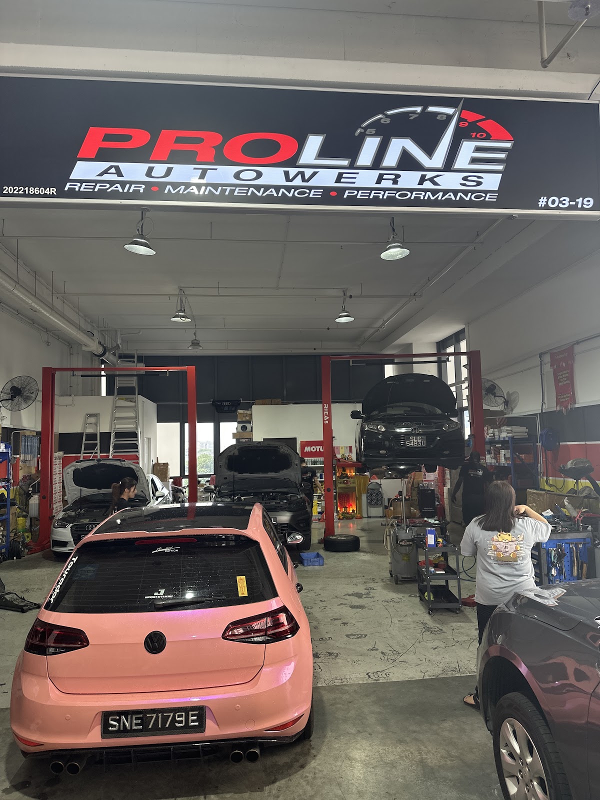 The illuminated Proline Autowerks signboard reading Repair, Maintenance, Performance above a busy workshop floor with an Audi, a Honda on a lift and a pink Volkswagen Golf